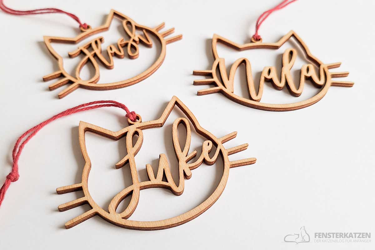 Drei Weihnachtsbaumanhänger aus Holz in Form von Katzenköpfen, jeder mit einem anderen Namen - Flash, Nala und Luke - in Schreibschrift ausgeschnitten. Sie hängen an roten Schnüren auf weißem Hintergrund und sind bezaubernde Geschenkideen, die Katzenliebhaber begeistern werden.